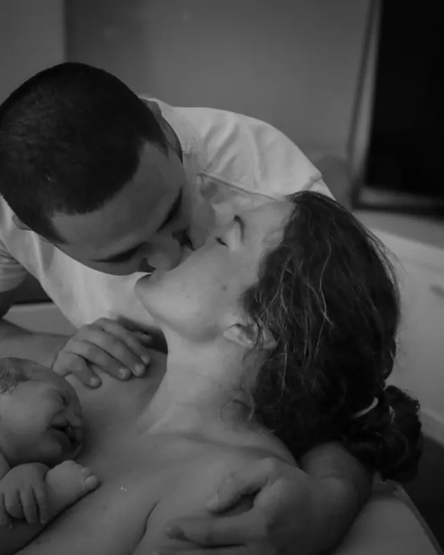 A father kisses mom as she holds their newborn, black and white birth photography by Jenn Miller, Orange County