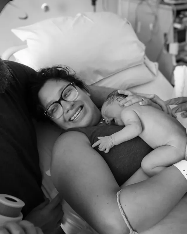 A mother beams with her newborn on her chest, black and white birth photography by Jenn Miller, Orange County