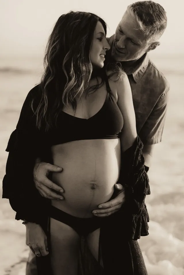 Jenn Miller, pregnant, held by her husband on the beach, in black and white