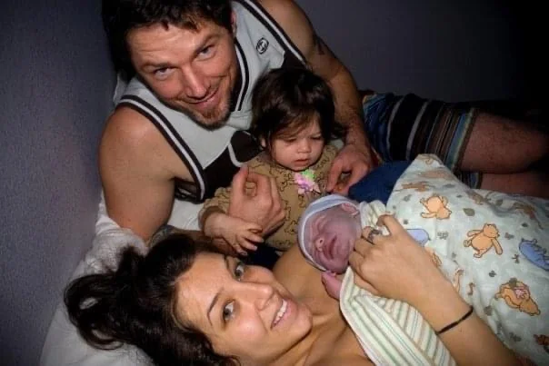 Jenn Miller smiling beside her swaddled newborn while her husband and toddler look on